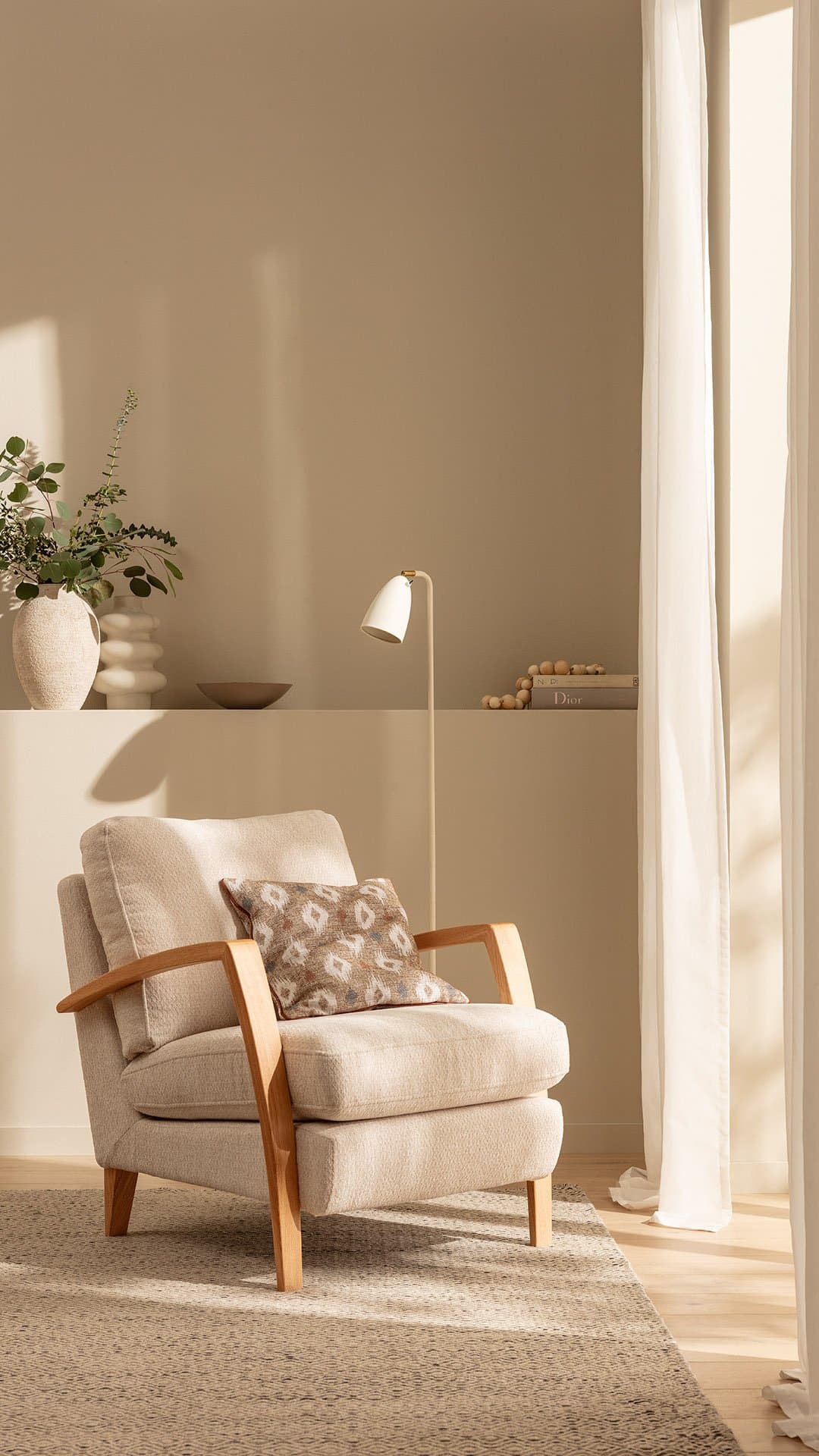 Beiger Sessel mit Holzarmlehnen, dekorativem Kissen und Stehlampe in einem hellen, minimalistischen Raum.