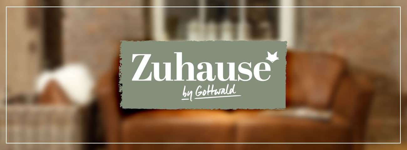 Logo mit dem Schriftzug "Zuhause by Goldblatt" auf unscharfem Hintergrund mit einer gemütlichen Sitzgelegenheit.