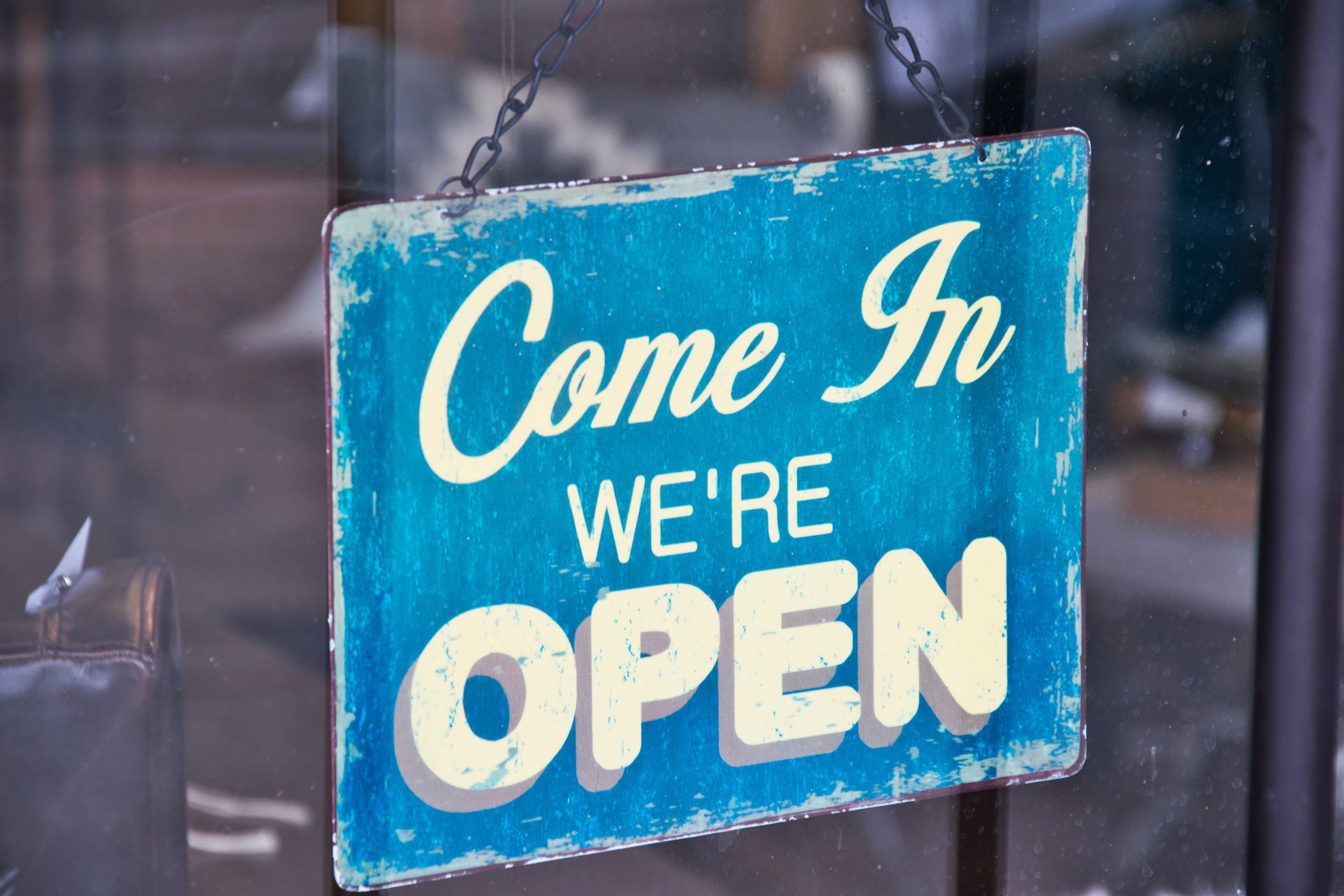 Türschild „Come In – We're Open“ an einer Glastür