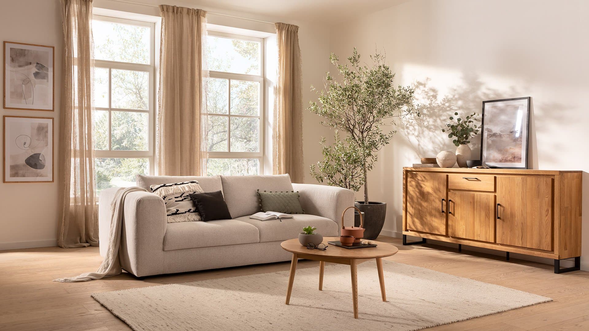Helles Wohnzimmer mit hellbeiger Couch, Holzsideboard, runden Tisch und Pflanzen, durch große Fenster beleuchtet.