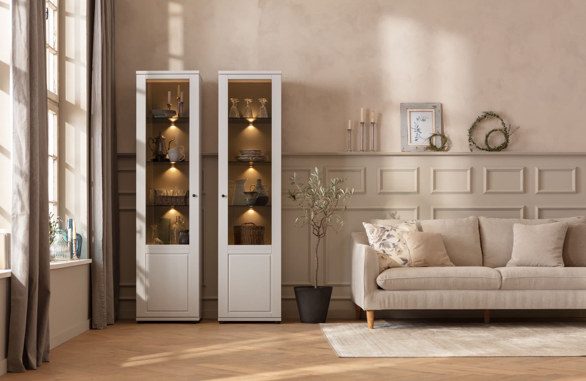 Heller Wohnraum mit einem beigen Sofa, zwei weißen Vitrinen und einer sanften Lichtgebung, umgeben von eleganten Deko-Elementen.