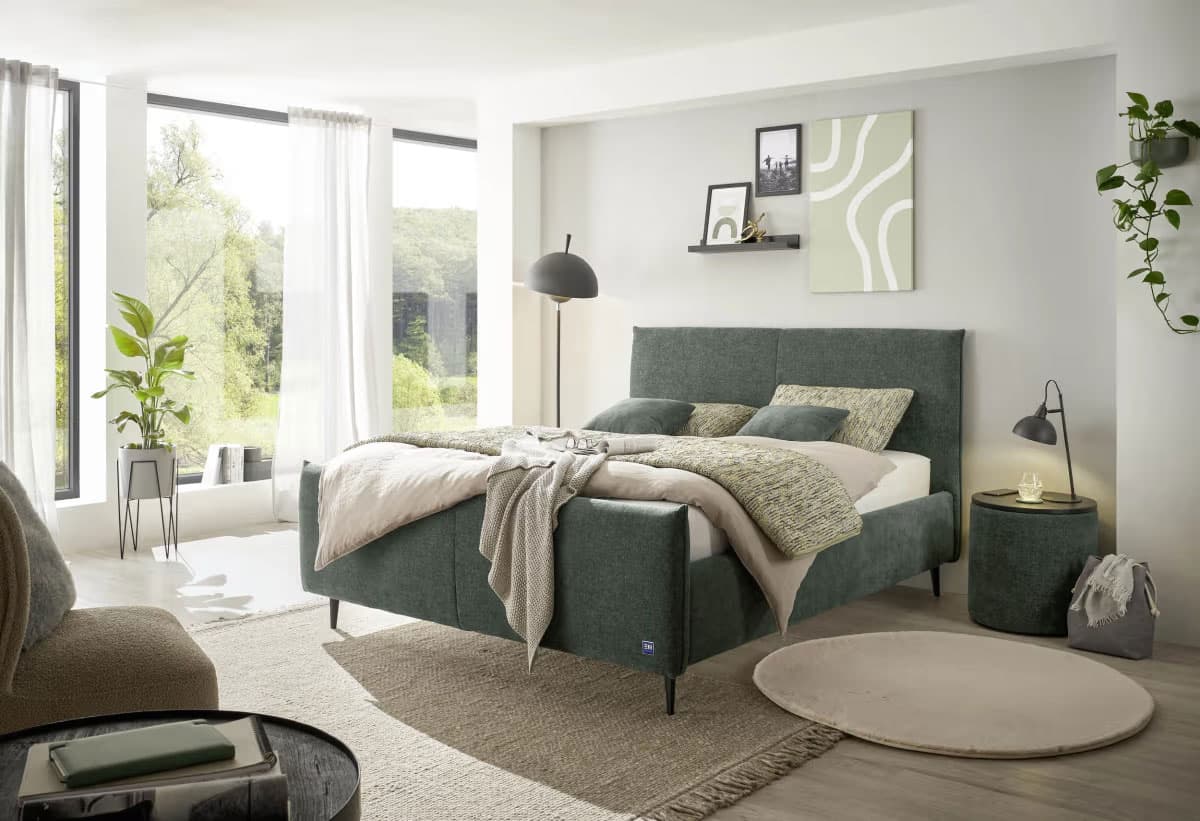 Modernes Schlafzimmer mit grünem Bett, sanften Textilien und natürlicher Beleuchtung durch große Fenster. Pflanzen und kreative Wanddekoration.