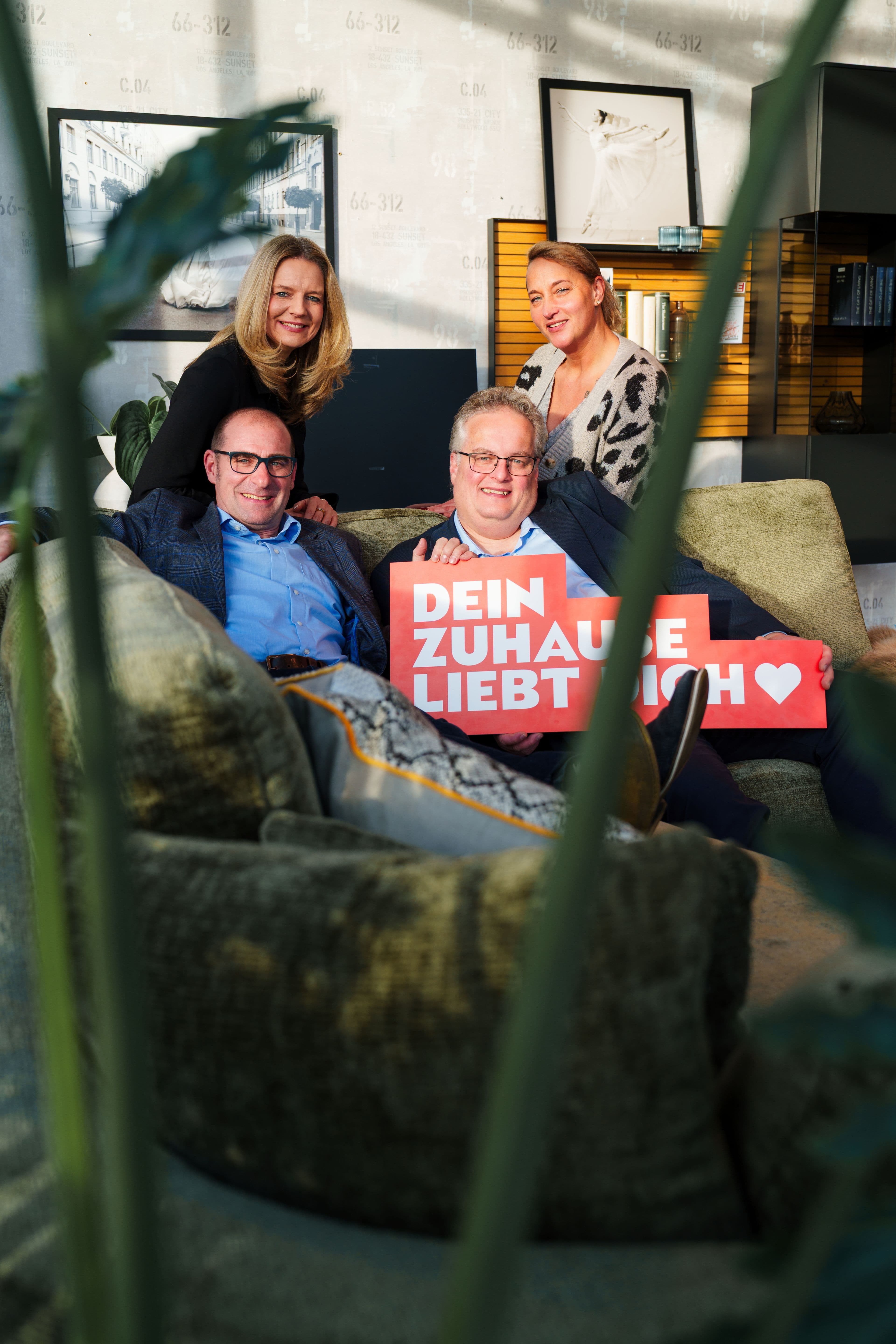 Vier Personen sitzen auf einem Sofa in einem modernen Wohnzimmer und halten ein Schild mit der Aufschrift „DEIN ZUHAUSE LIEBT DICH“.