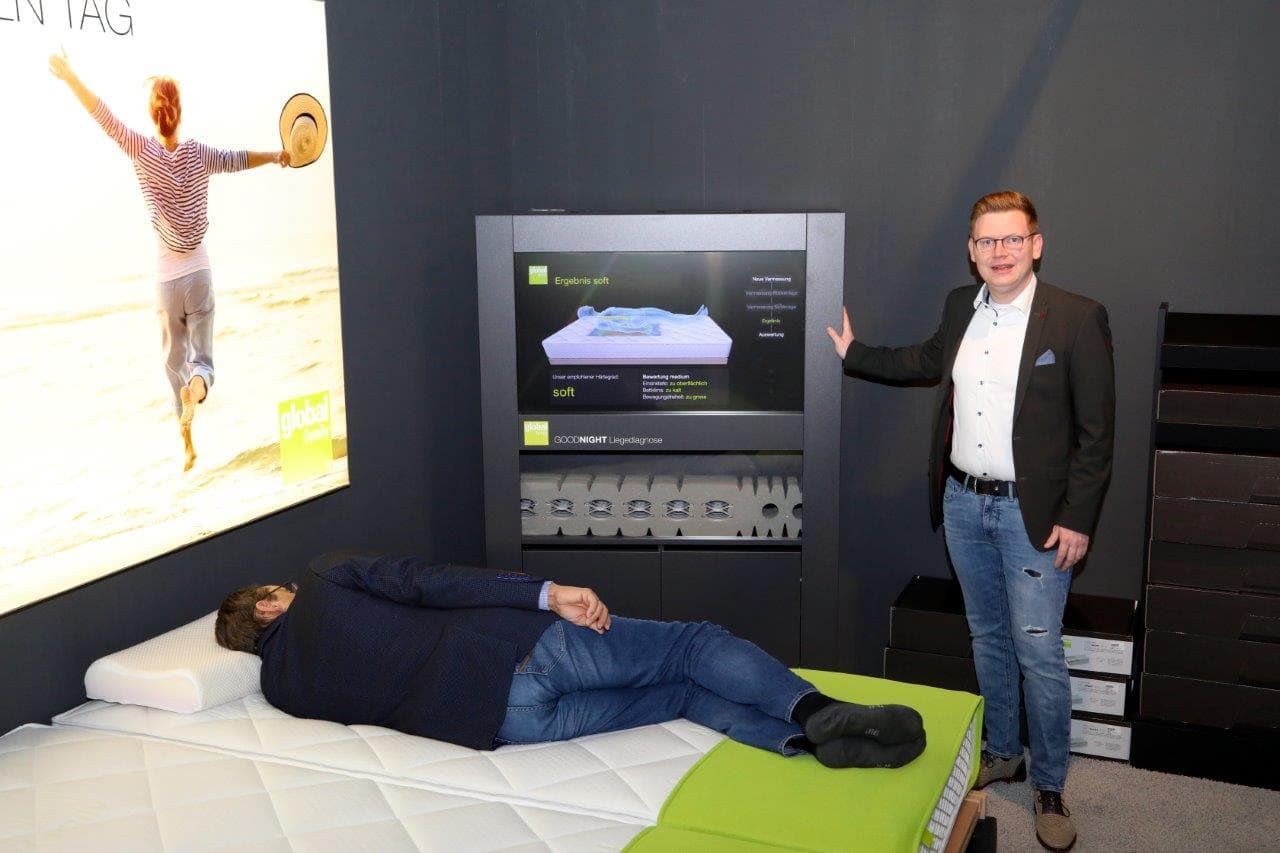 Ein Mann steht neben einem Bildschirm, während ein anderer Mann auf einer Matratze liegt. Hintergrund ist ein großes Werbebild.