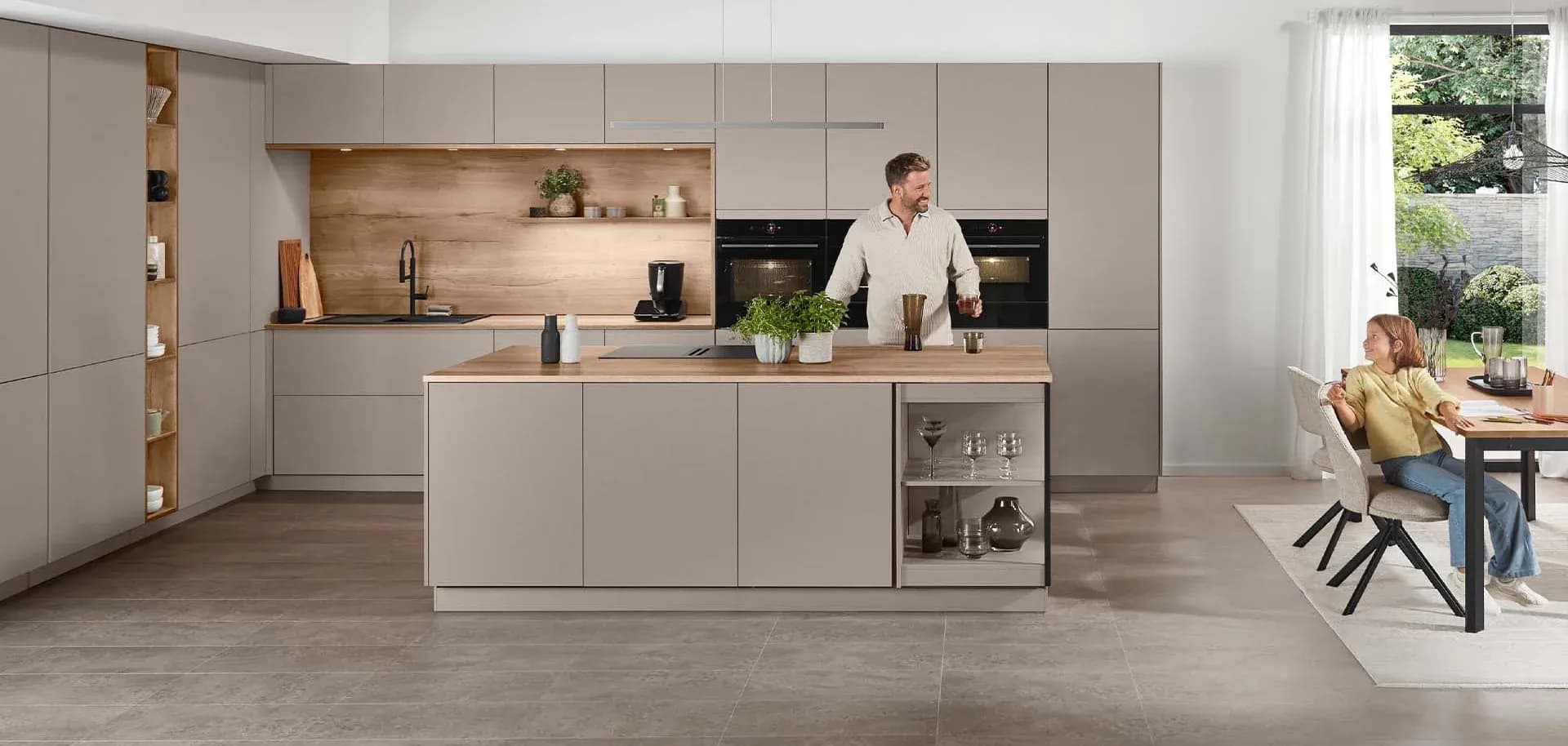 Moderne, helle Küche mit Kochinsel, Holzregal und Tisch, an dem eine Person sitzt und eine andere kocht.