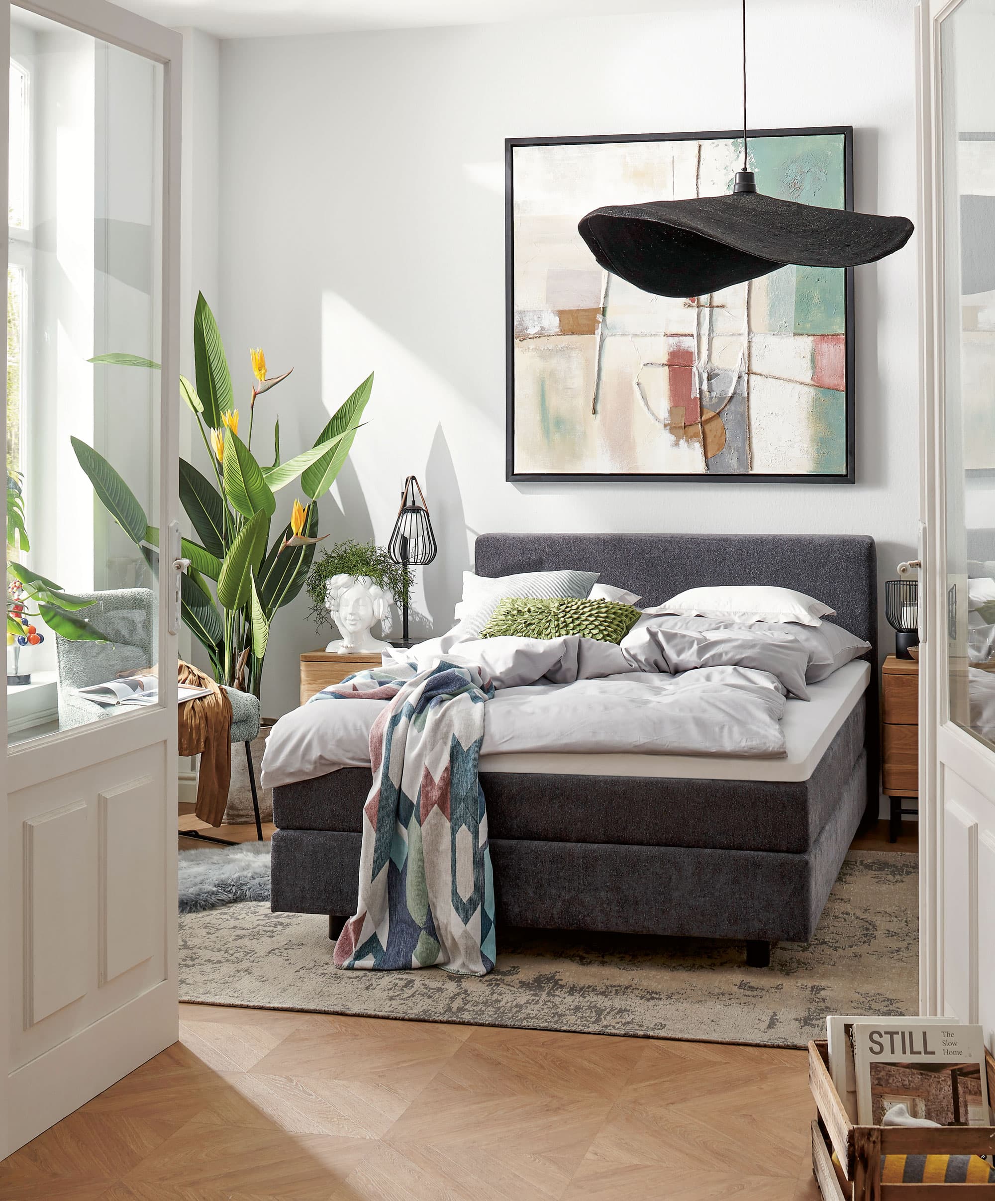 Helles Schlafzimmer mit einem großen Bett, modernen Möbeln und einer Pflanze im Hintergrund. Dekoratives Gemälde an der Wand.