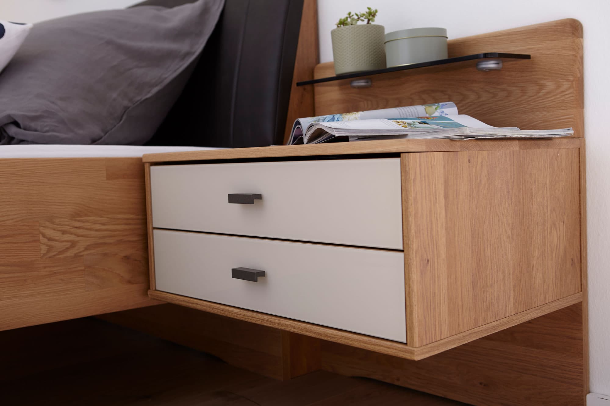 Nachttisch aus Holz mit zwei Schubladen, einer Glasablage und einem Pflanzengefäß, neben einem Bett mit grauem Kopfteil.
