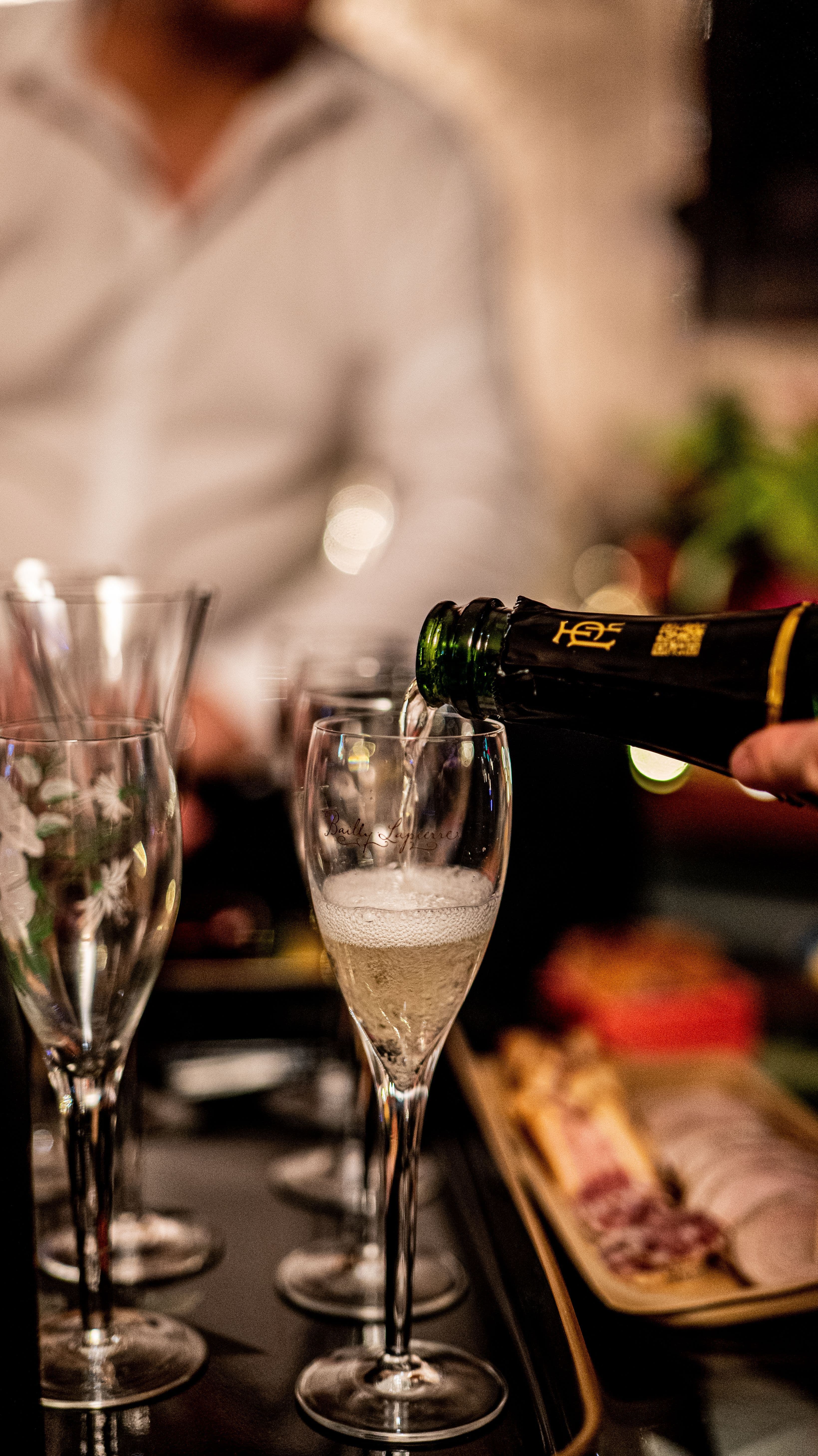 Sekt wird in ein Glas eingeschenkt, im Hintergrund sind weitere Gläser und eine Auswahl an Snacks zu sehen.
